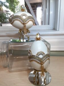 Two jeweled eggs. One at the bottom on top of a reflective sliver stands, the other sitting atop a glass jar.
